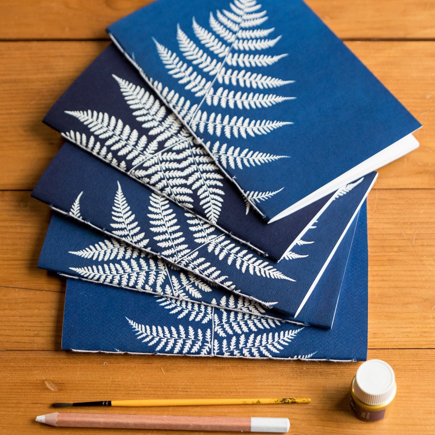 Blue notebooks with white fern patterns on a wooden surface with paintbrushes and a bottle.