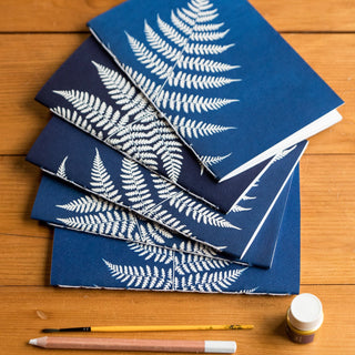 Blue notebooks with white fern patterns on a wooden surface with paintbrushes and a bottle.