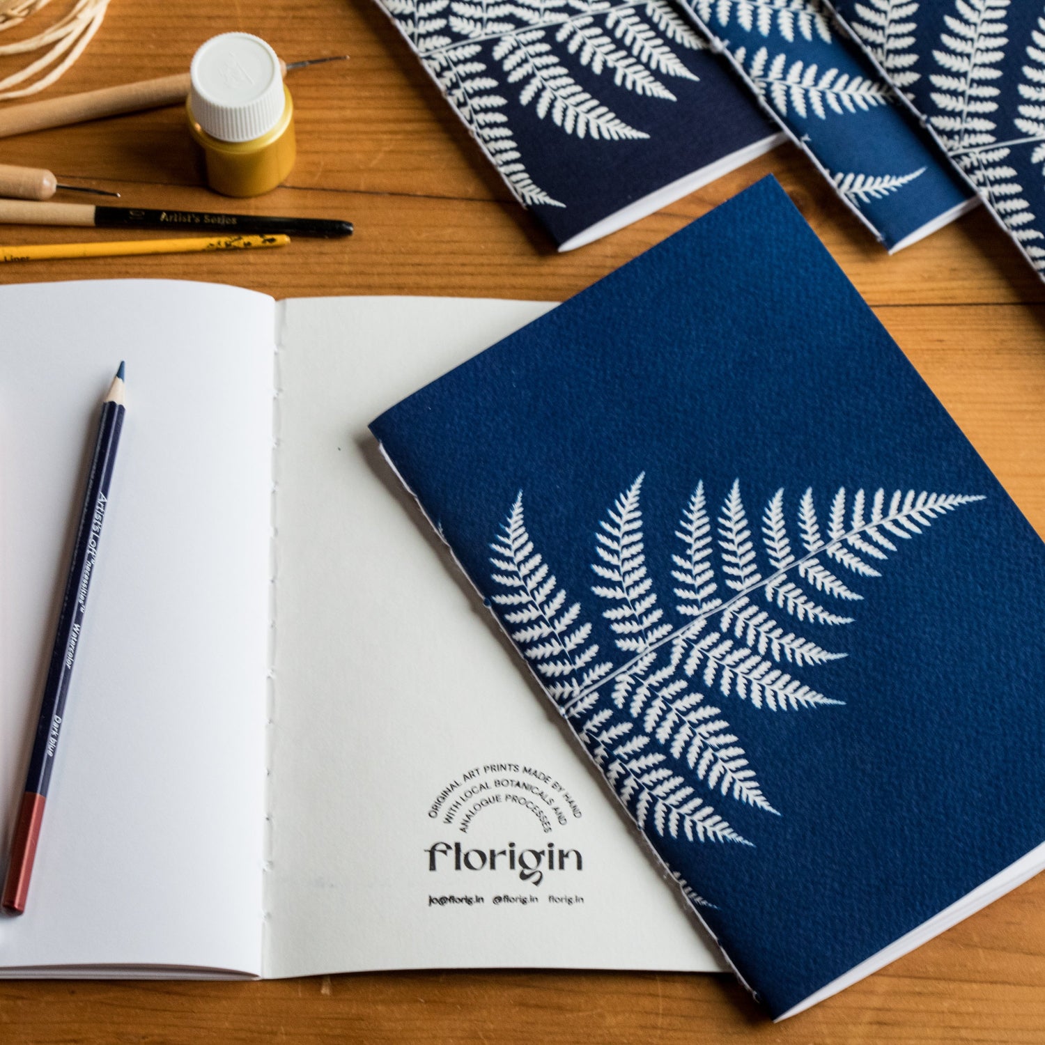 Notebook with white leaf design on blue cover, open book with pens, and paint supplies on a wooden surface.