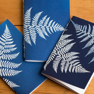 Three blue notebooks with white leaf patterns on a wooden surface, accompanied by two paintbrushes.