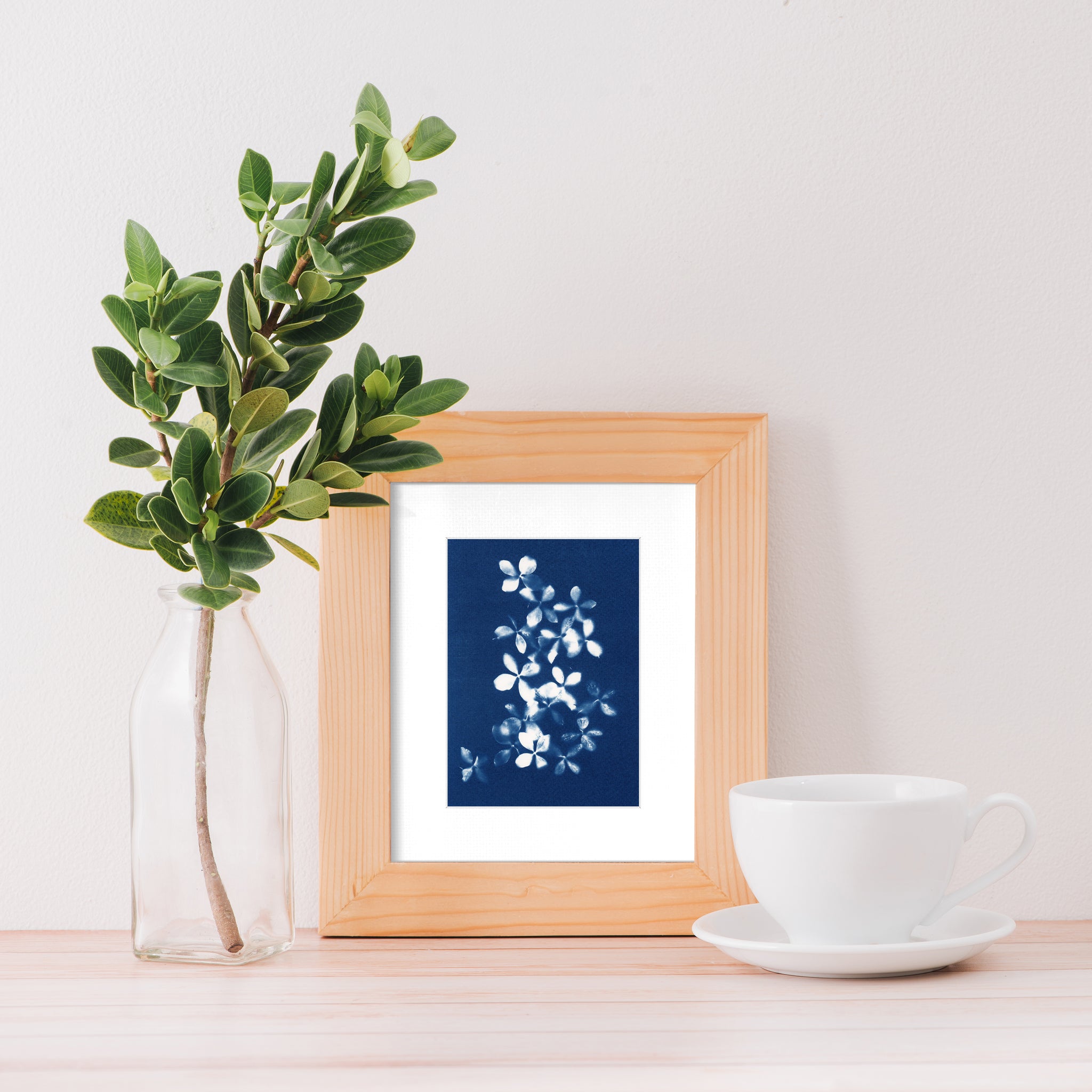 frame with blue cyanotype art print by Jo Thomson on a side table with coffee cup and leaf stem in a bottle vase